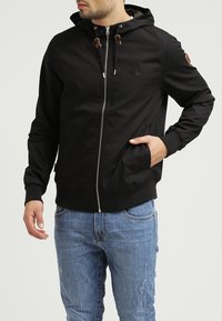 Czarna kurtka z kapturkiem, zamkiem błyskawicznym, bocznymi kieszeniami i ściągaczami na rękawach. Posiada brązowe akcenty ze sznurka oraz naszywkę z logo na rękawie.