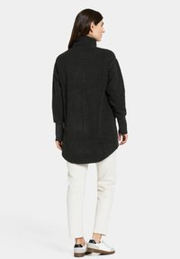 Schwarzes, strukturiertes Fleece-Oberteil mit langen Ärmeln, hohem Kragen und abgerundetem Saum, kombiniert mit hellen Hosen und weißen Sneakern.