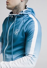 Blå gradient lynlås-hættetrøje med hvidt logo, kontrasterende hvide sidestriber og snørret hætte. Stoffet virker glat med let glans.