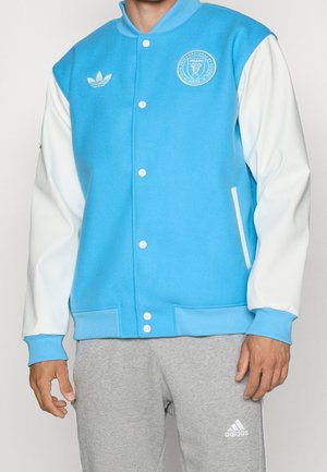 Hombre que lleva una chaqueta universitaria azul y blanca con el logo de Miami y pantalones deportivos grises de Adidas con logo en el muslo izquierdo.