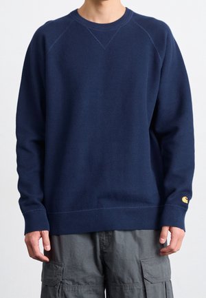 Homme portant un pull bleu marine à manches longues avec poignets et ourlet côtelés, associé à un pantalon cargo gris sur un fond uni.