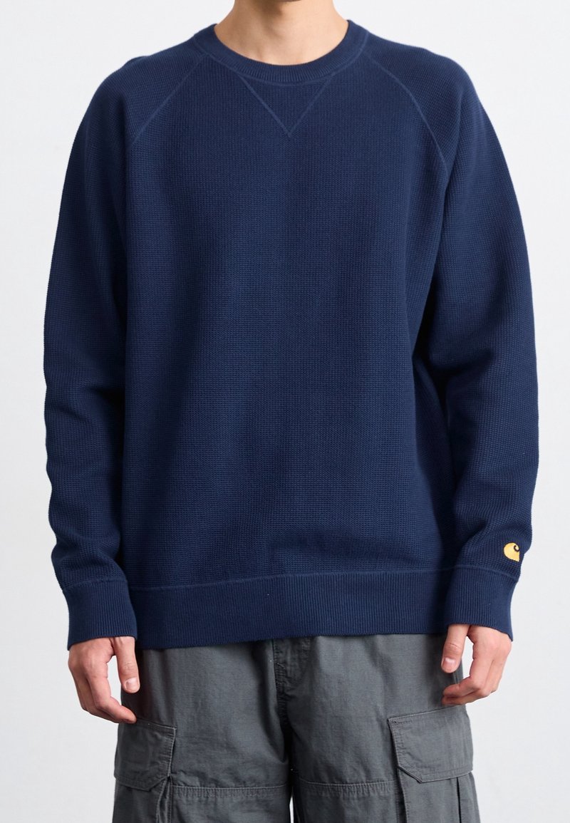 Homme portant un pull bleu marine à manches longues avec poignets et ourlet côtelés, associé à un pantalon cargo gris sur un fond uni.