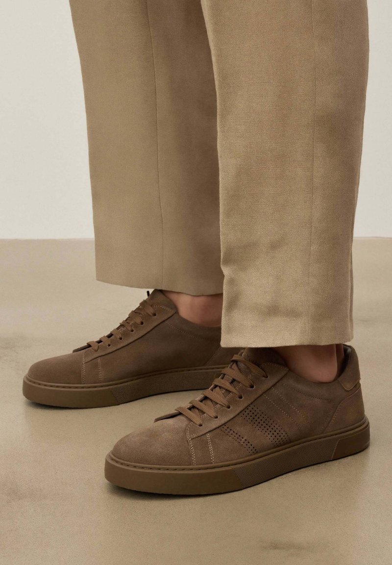 Baskets en daim marron à lacets portées avec un pantalon beige sur un sol et un fond neutres.