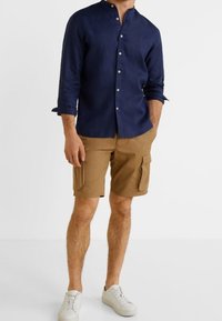 Marineblauw overhemd met knopen en een opstaande kraag, gecombineerd met khaki cargoshorts en witte sneakers. Het overhemd heeft lange mouwen die bij de manchetten zijn opgerold.