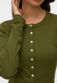 Žena nosi maslinasto zelenu rebrastu majicu dugih rukava s okomitim redom bijelih dugmadi sprijeda, ruka joj je blizu lica.