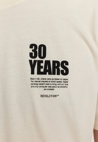 T-shirt en coton couleur crème avec un texte noir audacieux indiquant "30 ANS" et d'autres détails imprimés. Design à col rond, manches courtes.