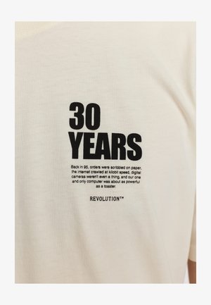 Crème-kleurige katoenen t-shirt met vetgedrukte zwarte tekst "30 JAAR" en extra bedrukte details. Crewneck ontwerp, korte mouwen.