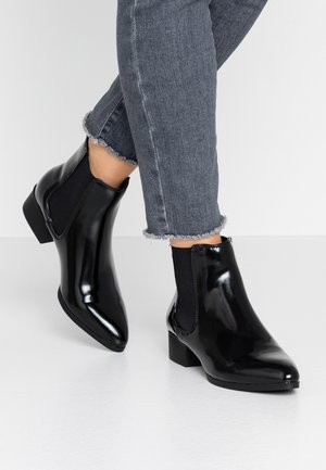 Bottes cheville noires brillantes avec des panneaux latéraux en élastique, un bout pointu et un talon bloc bas. Portées avec un jean denim gris coupé court.