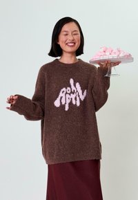 Rjav puhast pulover z belim grafičnim dizajnom, v kombinaciji z maroon krilom. Model drži stekleno krožnik s pink in belimi penicami.