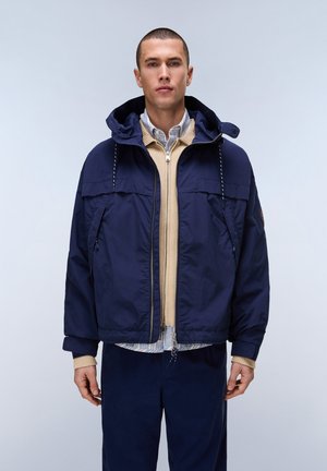 Mężczyzna ubrany warstwowo, w granatową kurtkę z kapturem, beżowy sweter na zamek i pasiastą koszulę z kołnierzykiem, stojący na jednolitym tle.