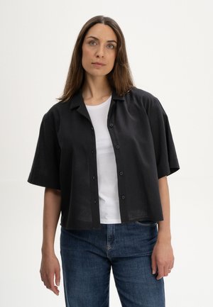 Vrouw met lang bruin haar die een zwart, kort mouwtjes, knoopshirt draagt over een wit topje en blauwe spijkerbroek, staand tegen een effen achtergrond.