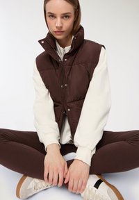 Gilet imbottito marrone con colletto alto, bottoni a pressione argentati e zip. Indossato sopra un maglione crema, abbinato a leggings marroni e sneakers bianche.