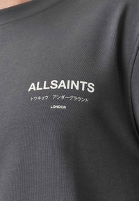Γκρι ύφασμα με λευκό κείμενο που γράφει "ALLSAINTS" με μεγάλα γράμματα, ιαπωνικούς χαρακτήρες από κάτω και "LONDON" από κάτω τους.