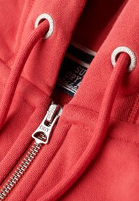 Rode hoodie met een zilveren rits en trekkoord, met metalen oogjes en een label dat "Superdry Athletic" leest. Glad textuur.