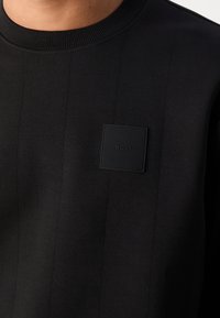 Gros plan sur un haut noir à col rond avec des coutures verticales et un petit patch noir texturé embossé du mot « BOSS » sur la poitrine.