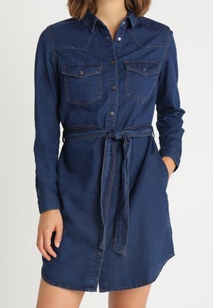 Langærmet mørkeblå denimskjortekjole med knapper foran, brystlommer og bundet bælte i taljen, båret af en stående person med den ene hånd i lommen.
