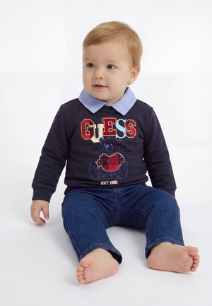 Jeune enfant aux cheveux clairs assis pieds nus, portant un sweat bleu marine avec un motif "GUESS" et un ours en peluche, ainsi qu'un jean bleu sur fond blanc.