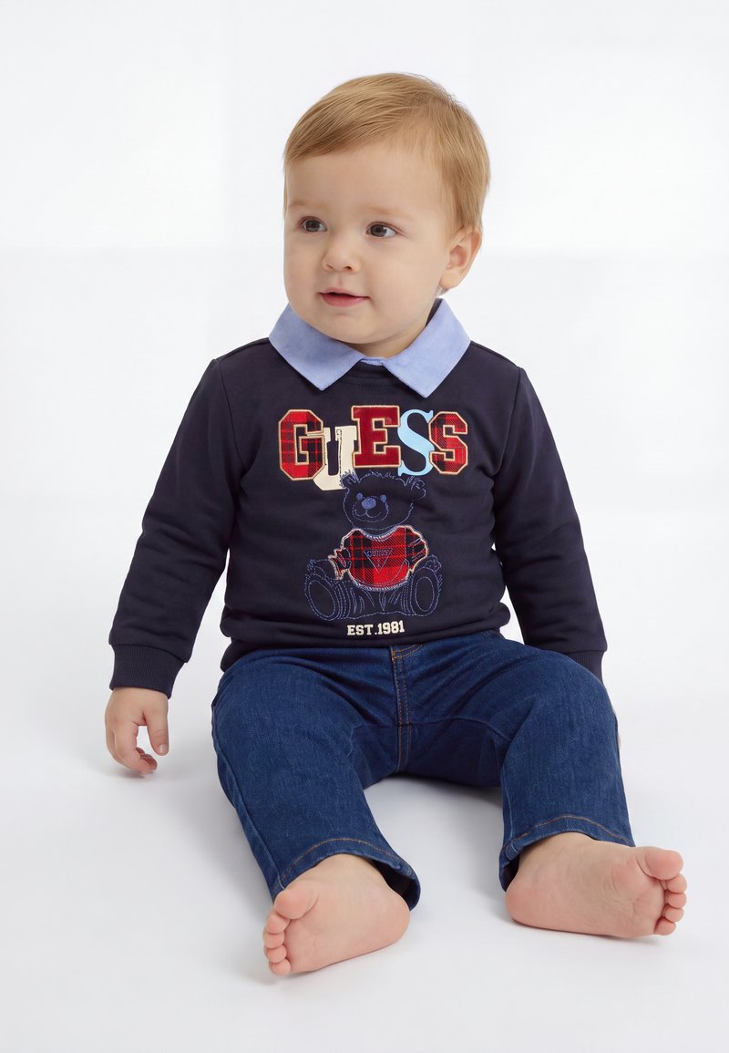 Jeune enfant aux cheveux clairs assis pieds nus, portant un sweat bleu marine avec un motif "GUESS" et un ours en peluche, ainsi qu'un jean bleu sur fond blanc.