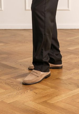 Feet wearing beige suede loafers paired with black pants, standing on a polished wooden parquet floor in an indoor setting.