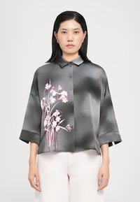 Camicia in satin grigia con un motivo floreale rosa, caratterizzata da una chiusura a bottoni sul davanti, un colletto e maniche larghe, abbinata a pantaloni chiari.