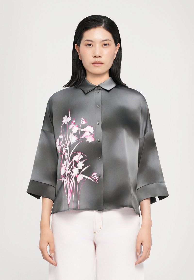 Camicia in satin grigia con un motivo floreale rosa, caratterizzata da una chiusura a bottoni sul davanti, un colletto e maniche larghe, abbinata a pantaloni chiari.
