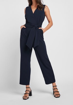 Femme portant une combinaison sans manches bleu marine avec une large ceinture à nouer et des sandales noires à talons blocs et à brides, debout devant un fond clair uni.