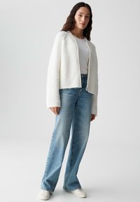 Witte fluffy cropped jas over een wit T-shirt, gecombineerd met lichtblauwe wijde spijkerbroek en witte sneakers. Soepele texturen, ontspannen pasvorm.