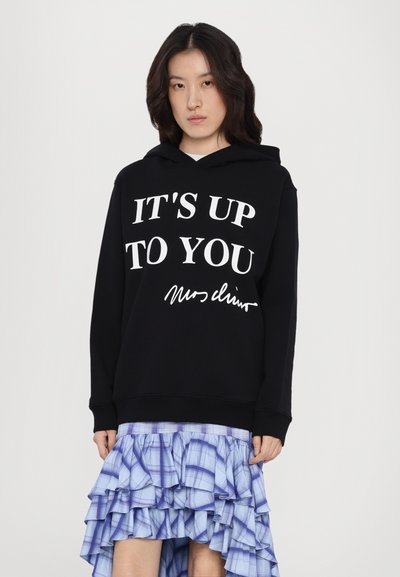 Svart hoodie med vit text "DET ÄR UPP TILL DIG" och signatur. Kombinerad med en ljusblå, veckad kjol med rutmönster.