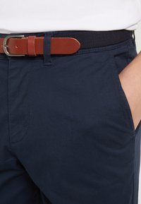 Person in einer dunkelblauen Hose mit einem braunen Ledergürtel, die rechte Hand liegt in der vorderen Tasche.