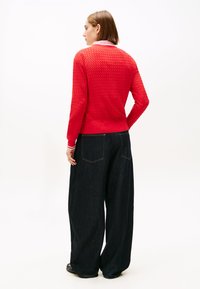 Κόκκινο πλεκτό πουλόβερ με υφή και στρογγυλή λαιμόκοψη, συνδυασμένο με παντελόνι από σκούρο τζιν φάρδους, που διαθέτει κλασικές πίσω τσέπες και ραφές λεπτομέρειας.