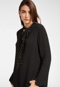 Blusa nera con maniche lunghe, decorata con pois dorati e chiusura a fiocco sul colletto. Presenta un orlo arrotondato e dettagli con pietre preziose.