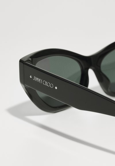 Black cat-eye sunglasses with dark green lenses. Features include a glossy finish and silver-accented logo on the arm.