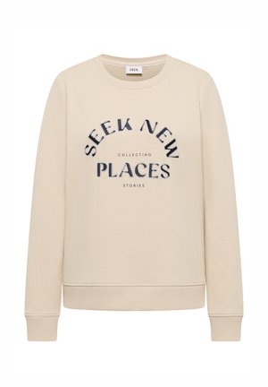 Beżowa bluza z prążkowanymi mankietami i ściągaczem u dołu, z czarnym napisem: "SEEK NEW PLACES" oraz mniejszymi słowami "COLLECTING STORIES" poniżej.