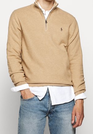 Man draagt een beige trui met kwart-ritssluiting over een wit overhemd met opgerolde manchetten en lichtblauwe spijkerbroek, hand in de zak.