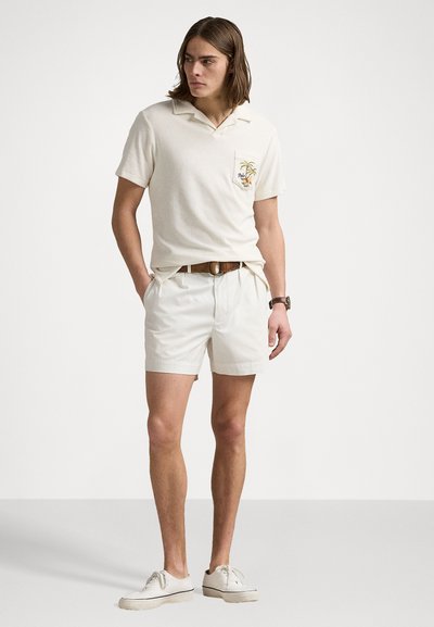 Bež majica s kratkimi rokavi, polo stil, s žepom in vezenjem palmovih dreves, v kombinaciji z belimi kratkimi hlačami in belimi čevlji.
