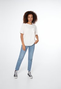 Crème-kleurige t-shirt met korte mouwen en een zak, lichtblauwe rechte jeans, en zwarte high-top sneakers tegen een witte achtergrond.