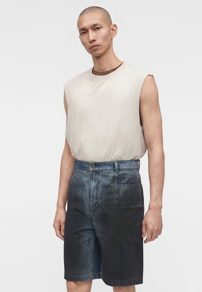 Άνδρας μοντέλο με ξυρισμένο κεφάλι φορώντας μανίκες αμάνικο τοπ σε εκρού χρώμα και μπλε τζιν σορτς με πολλαπλές τσέπες, στέκεται μπροστά σε απλό φόντο.