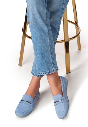 Blauwe suède loafers met een afgeronde neus, voorzien van een zilveren accent aan de voorkant, gedragen met opgerolde lichtblauwe jeans, zittend op een barkruk.