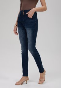 Mørk vask skinny jeans med høy midje, tre-knapp lukking og klassisk fem-lomme design. Glatt denim tekstur med lett falming.