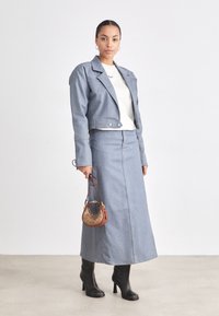 Set in denim composto da giacca e gonna di un blu chiaro, con spalle strutturate e dettagli in bottone, abbinato a una borsa marrone e stivali neri con tacco.