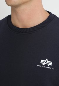 Maglione girocollo blu navy realizzato in tessuto liscio, con un patch bianco con il logo "Alpha Industries" sul petto. Collo a costine.