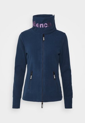Veste polaire bleu marine zippée avec col montant affichant le logo "Bench" en violet, avec des poches latérales zippées et des tirettes de fermeture éclair de marque.