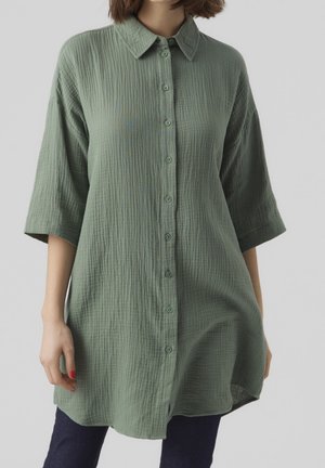 Vrouw draagt een losse, gestructureerde groene blouse met knopen en driekwartmouwen over een donkere broek, staand tegen een effen lichte achtergrond.