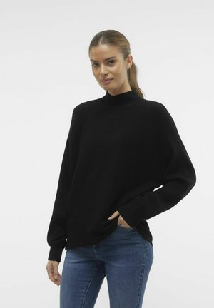 Maglione a coste nere con colletto alto e vestibilità ampia. Maniche lunghe e tasche laterali. Indossato sopra jeans in denim blu.
