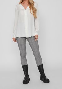 Blusa bianca a maniche lunghe con scollatura a V e chiusura a bottoni, abbinata a leggings grigi a quadri e stivale nero al ginocchio con zeppa.