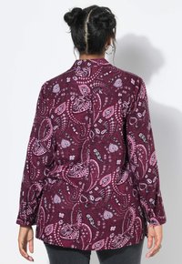 Vrouw met een gevlochten haarbun, draagt een langemouw shirt met paarse paisleyprint, met haar rug naar de camera gekeerd tegen een witte achtergrond.