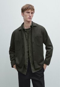 Giovane uomo con capelli corti che indossa una camicia verde scuro a bottoni sopra una t-shirt verde più scura e pantaloni neri, con le mani nelle tasche.