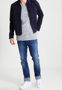 Blouson aviateur bleu marine, chemise à rayures grises, jean bleu et baskets blanches. Le blouson comporte deux poches avant et le jean a des revers aux poignets.
