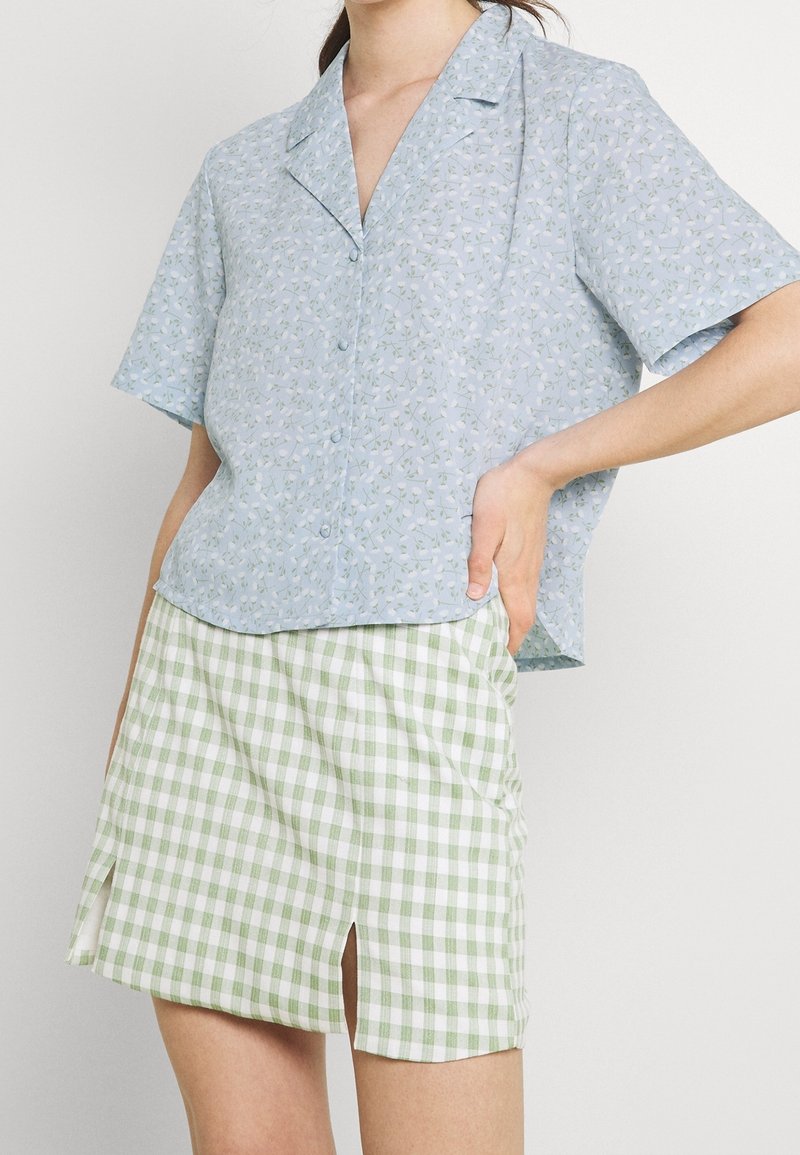 Camicia a maniche corte, azzurra, con bottoni e fantasia floreale; abbinata a una mini gonna a scacchi verde e bianca con spacchi laterali.
