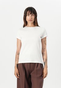 White cotton t-shirt with short rolled sleeves, rounded neck, and a straight hem. Worn with brown loose trousers. Model stands against a white background.